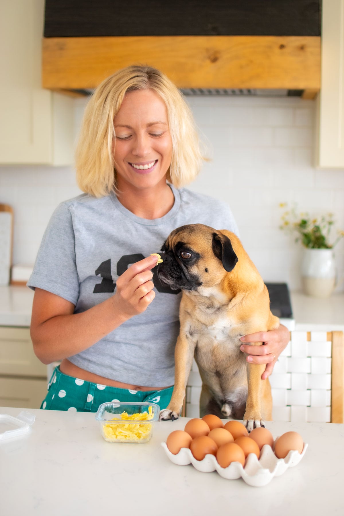 woman and pug in kitchen with a tupperware of scrambled eggs and a tray of brown eggs.