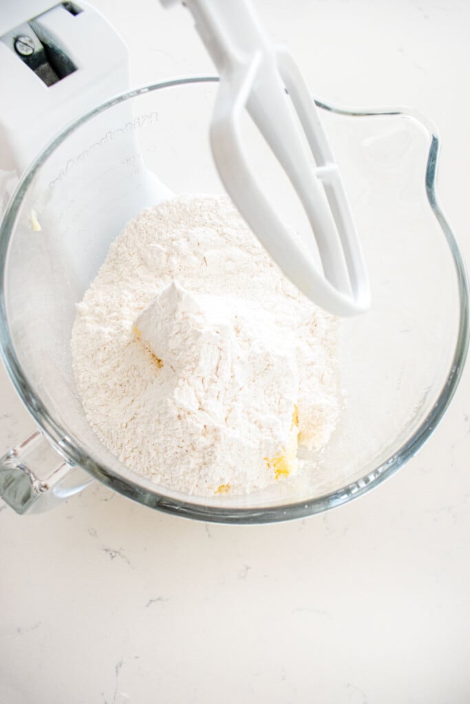 glass mixing bowl with powdered sugar, butter and vanilla extract on a marble counter.