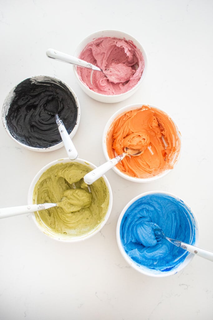 five bowls with different colored buttercream frosings on a marble counter.