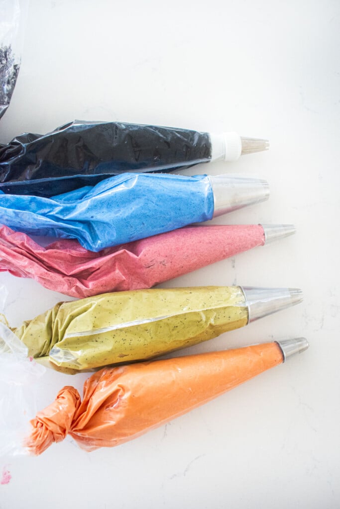 piping bags with different colored frostings and decorating tips on a marble counter.