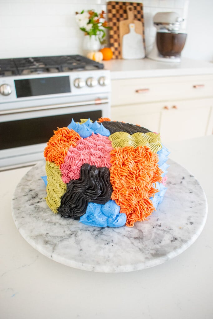monster cake piped with different colored frostings and textures on a marble turntable on the kitchen counter.