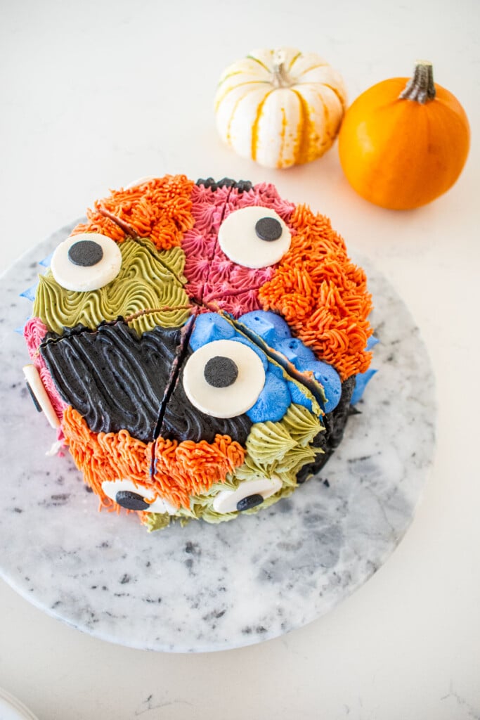 monster cake with different colored textures and big eyes on a marble turn table on a kitchen counter cut into triangle slices.